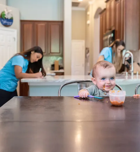 baby eating at a table while merry maids clean in the background