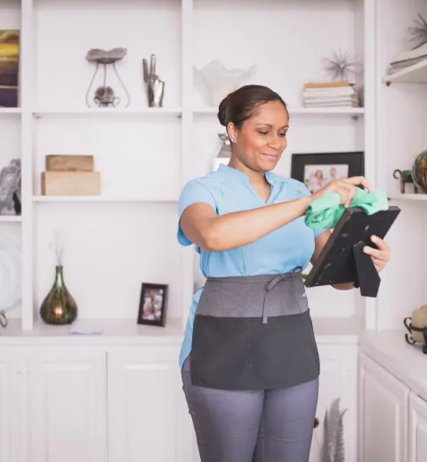 Merry Maids house cleaner carefully dusting picture frames and home decor on shelving.