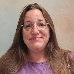 smiling woman with long brown hair standing in front of a grey wall wearing a purple shirt