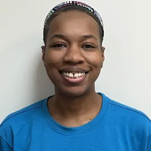 rayshell is smiling in front of a grey wall wearing a blue merry maids tshirt