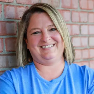 crystal has short blonde hair and is wearing a blue merry maids tshirt leaning against a brick wall