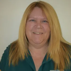 pam is wearing a green merry maids shirt standing in front of a tan wall