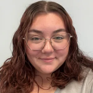 Gabby is wearing glasses, smiling in front of a tan wall