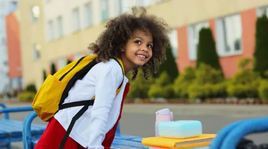 Young girl excitedly standing outside of an elementary school before the first day. She has her books next to her and is smiling by benches out front before starting her first day of a new grade. | Merry Maids®