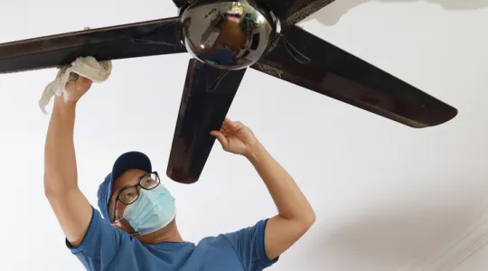 Low-angle view of an Asian man cleaning ceiling fan at home | Merry Maids
