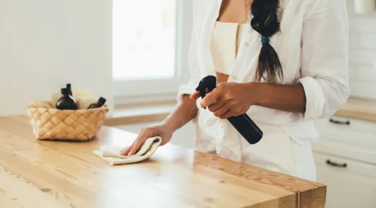 A young adult woman cleans her wooden kitchen island with a spray bottle and a rag. | Merry Maids