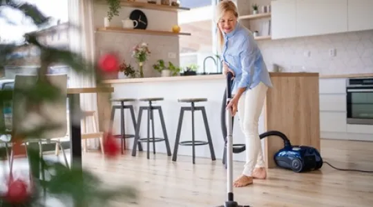 Woman smiling as she vacuums her hardwood floors in preparation of holiday company. She is tidying up and looking forward to her loved ones arriving. | Merry Maids®