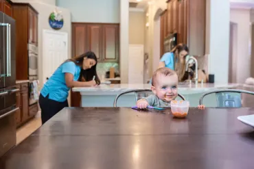 baby eating at a table while merry maids clean in the background