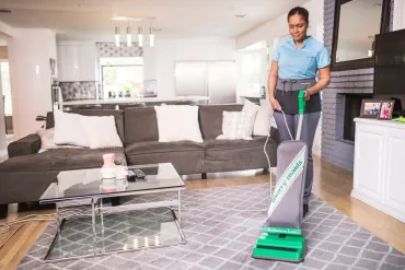 Merry Maids house cleaner vacuuming an area rug in a residential living room.